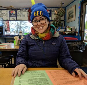 Grad student Ashli Anda sits in a restaurant before COVID and smiles at the camera; she is wearing a GEO beanie and a scarf and jacket