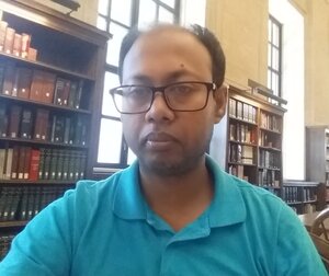Alamgir Hossain sits in front of two bookshelves and windows during day time.