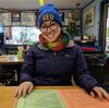 Grad student Ashli Anda sits in a restaurant before COVID and smiles at the camera; she is wearing a GEO beanie and a scarf and jacket