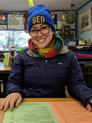 Grad student Ashli Anda sits in a restaurant before COVID and smiles at the camera; she is wearing a GEO beanie and a scarf and jacket
